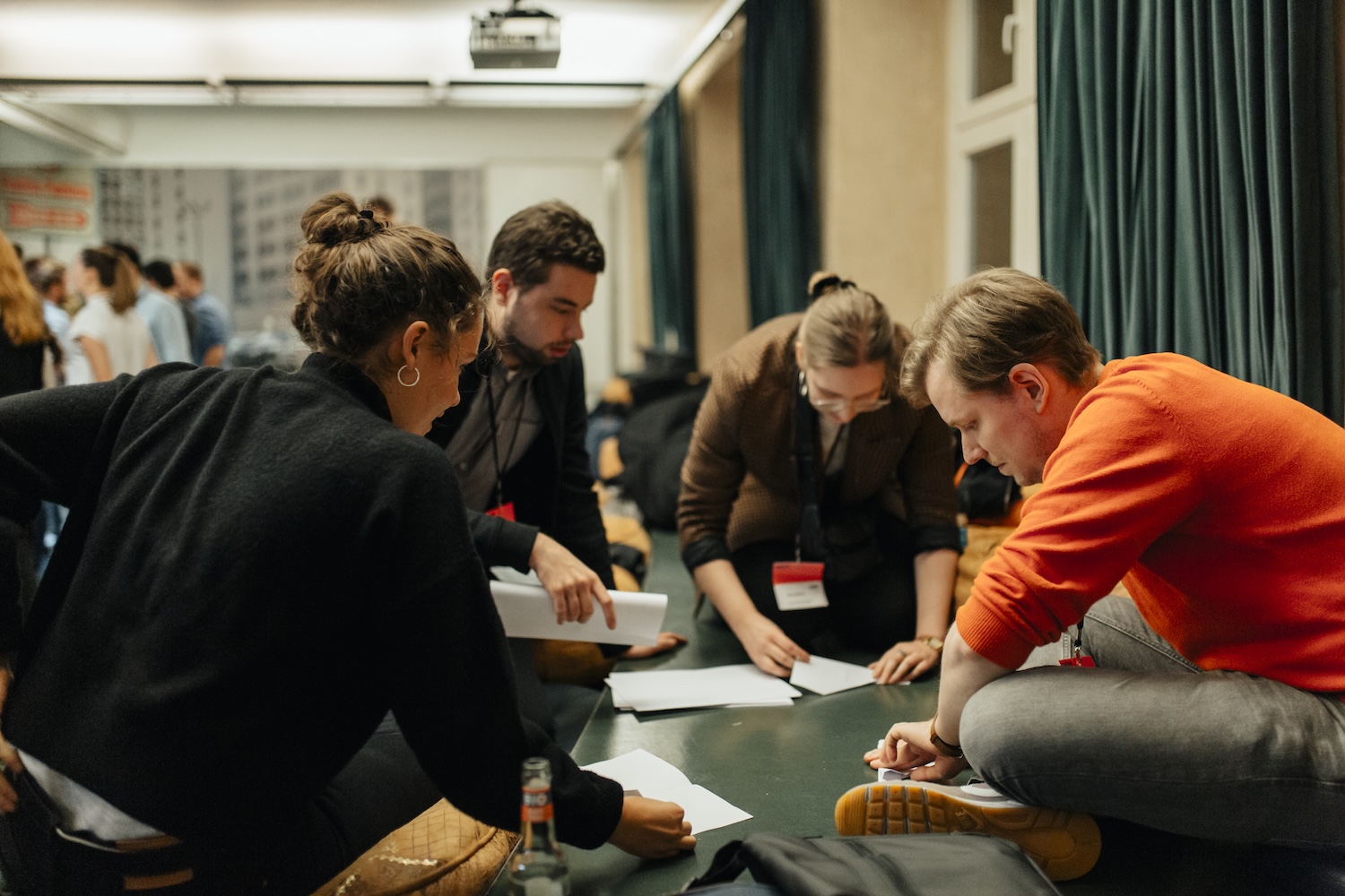 Vier Personen, zwei Männer und zwei Frauen, hocken um einen Tisch und arbeiten gemeinsam an Papieren. Sie sind konzentriert in einer Brainstorming-Session, mit weiteren Personen im Hintergrund des Raums. Auf dem Tisch steht eine Flasche, einige der Teilnehmer tragen Konferenz-Lanyards. nextMedia.Hamburg