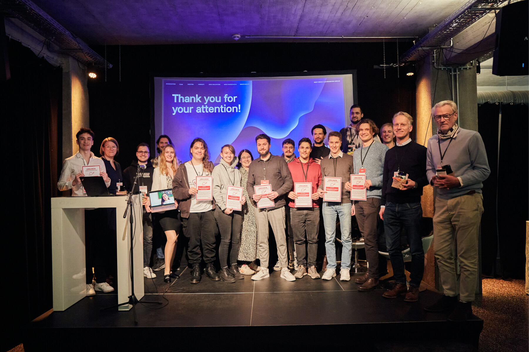 The group of 18 participants of the stands on a stage at the closing event of the Prototyping Lab 2022 and holds certificates in their hands. They smile at the camera while a large screen with the words 'Thank you for your attention! nextMedia.Hamburg