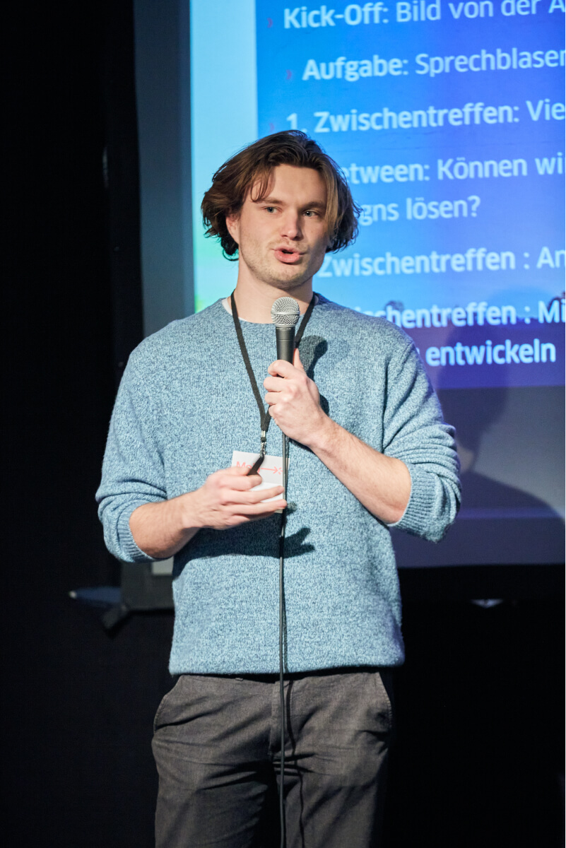 A young man with brown-blonde hair, representing Team Carlsen, holds a microphone and speaks on a stage. A projection with a presentation can be seen behind him. He is wearing a blue jumper, dark grey jeans and a conference lanyard. nextMedia.Hamburg