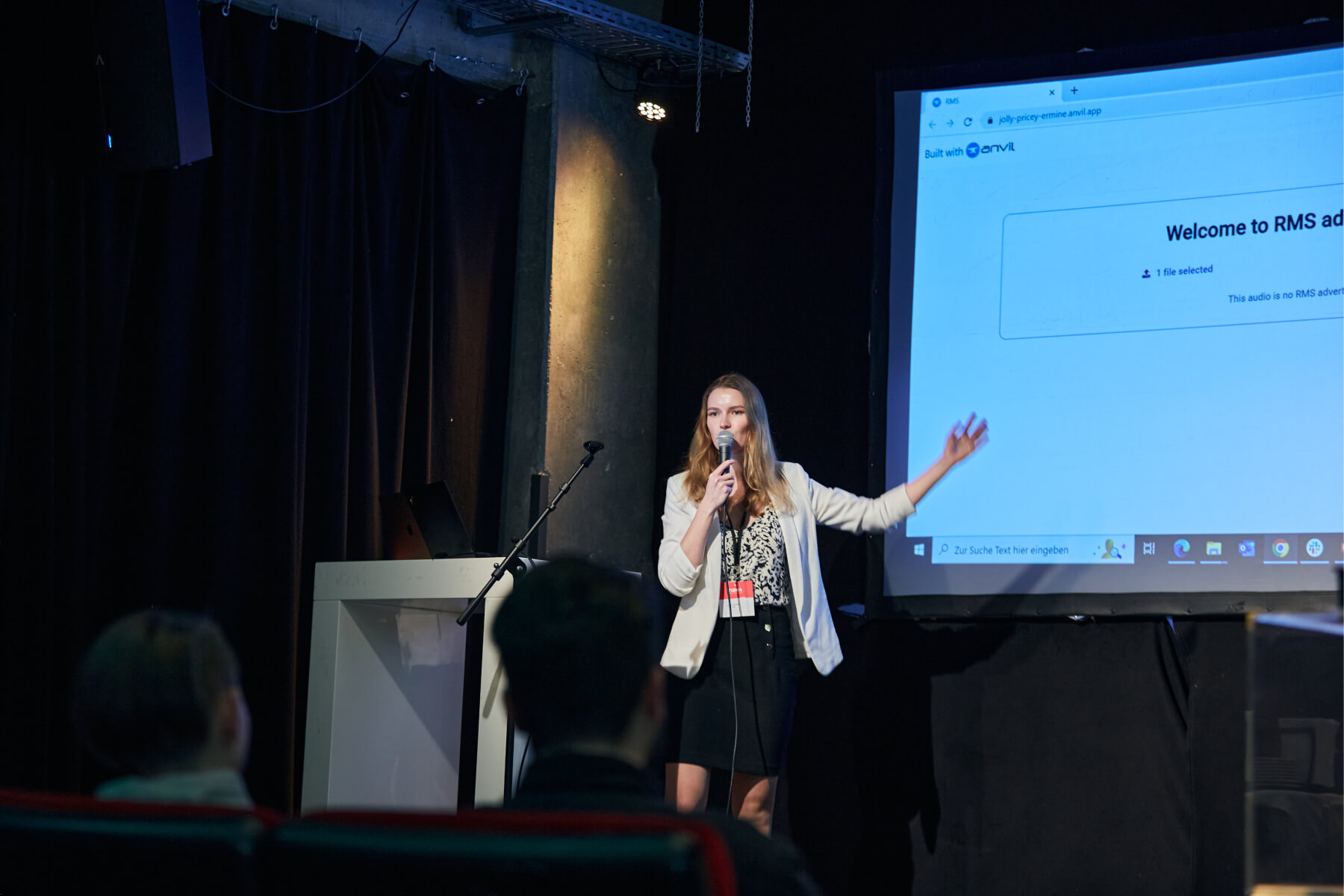 A young woman with dark blonde hair stands on a stage and speaks into a microphone. She is wearing a black skirt and a blouse with a floral black and white pattern and a white blazer over it. Two guests at the event can be seen blurred in the foreground. A projection of the Team RMS presentation can be seen in the background. nextMedia.Hamburg