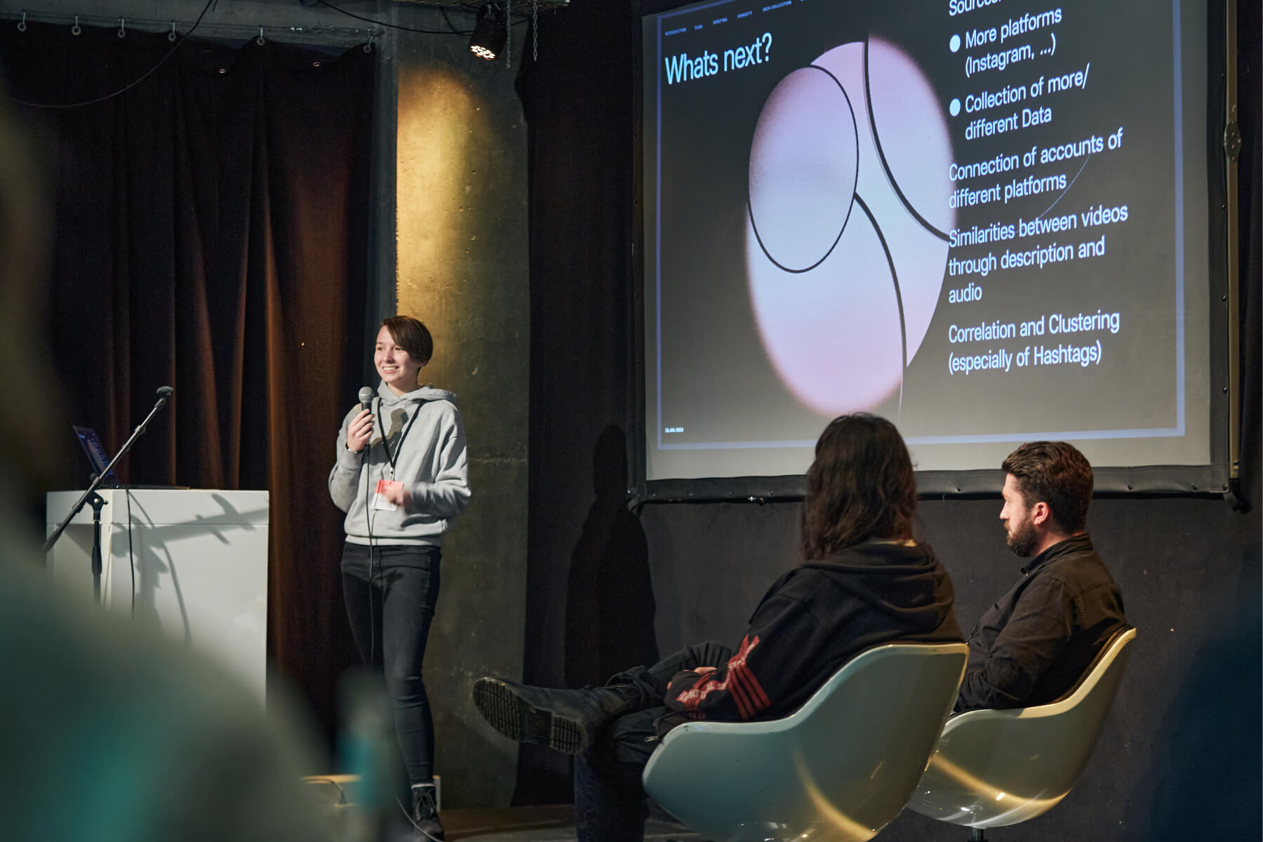 Presenter in a grey hoodie gives a talk on stage, behind her "What's next?" is shown on a projection screen. The two other members of the Jahreszeiten Verlag team sit in the foreground and listen attentively. nextMedia.Hamburg