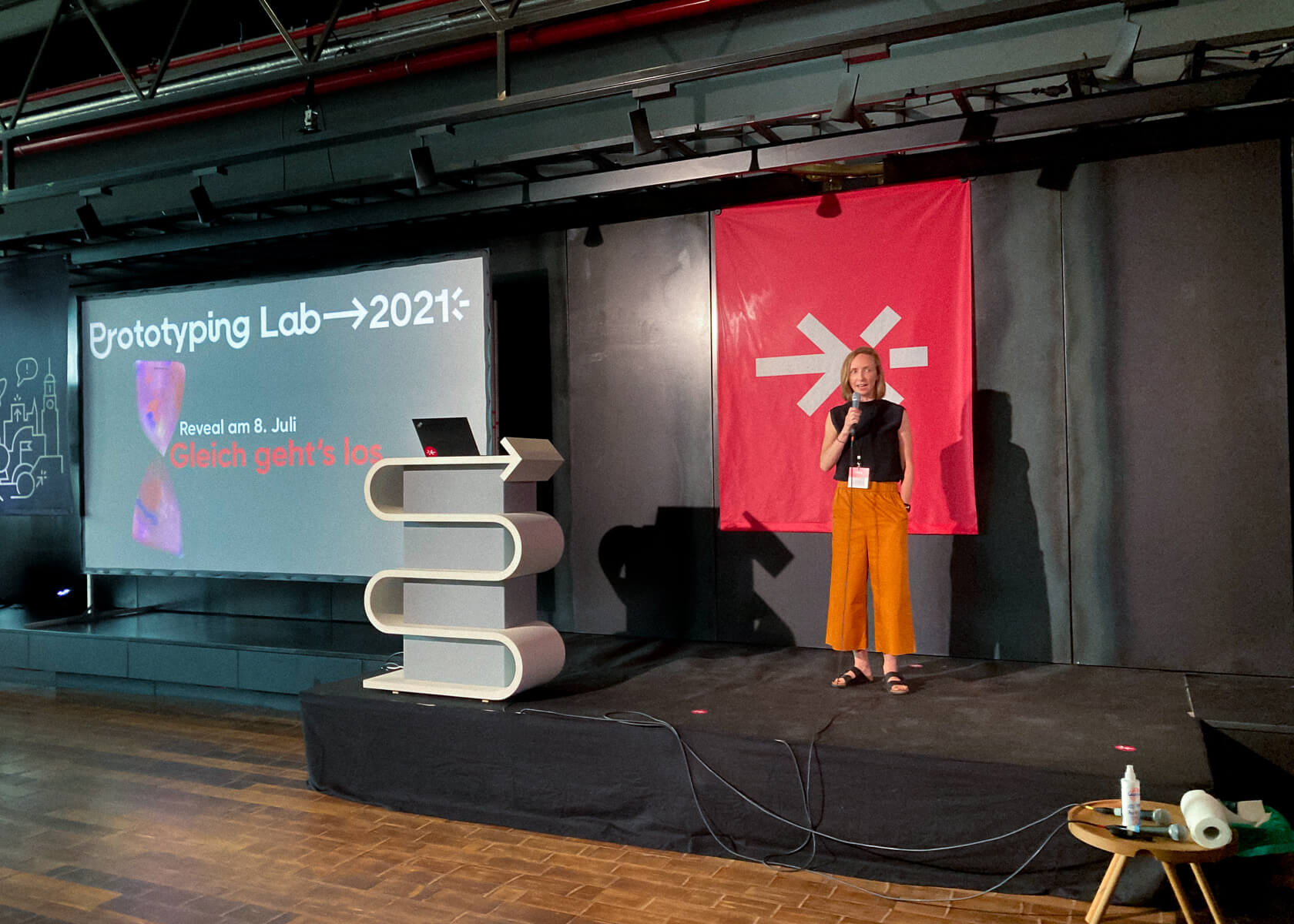 Event stage of the Prototyping Lab 2021 at the Design Centre Hamburg. The presenter in a black top and yellow trousers speaks at the microphone. The background shows a projected film with the words "Prototyping Lab 2021" and a red banner with the nextMedia arrow. nextMedia.Hamburg